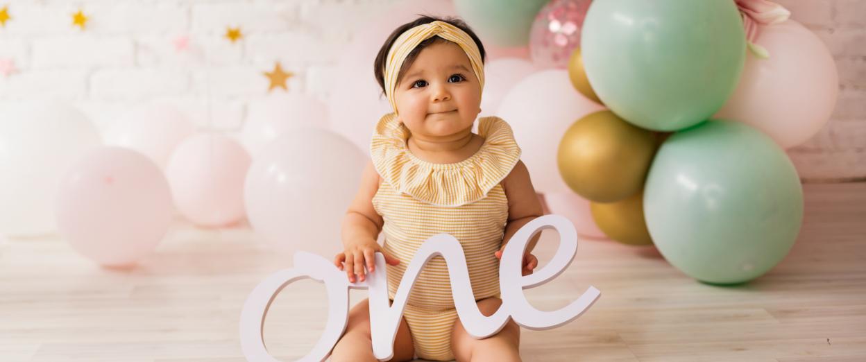 12 month old girl with olive complexion and brown hair in a yellow headband with birthday balloons behind her holding a sign that says "one". The balloons are light pink, light green, and gold