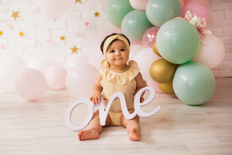 12 month old girl with olive complexion and brown hair in a yellow headband with birthday balloons behind her holding a sign that says "one". The balloons are light pink, light green, and gold