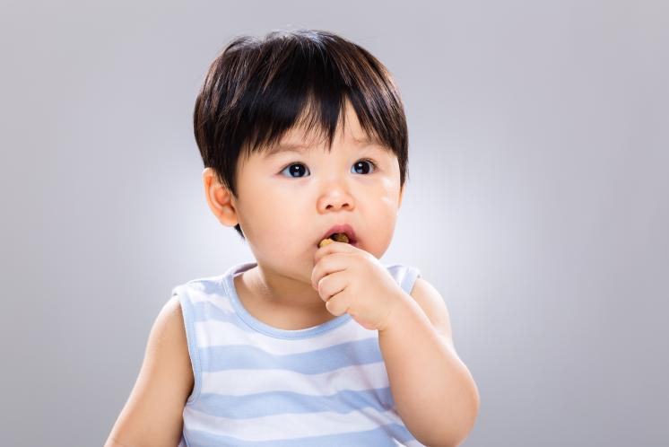 13 month old Asian boy with dark straight hair wearing a blue and white striped tank top looking at camera with something in his mouth he is taking a bite out of