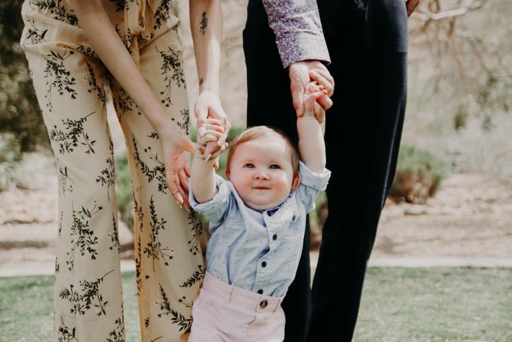 14 month old boy holding 2 adult's hands (the adults are cut off at the waist) while learning to walk. The baby has a light skin tone and reddish hair.