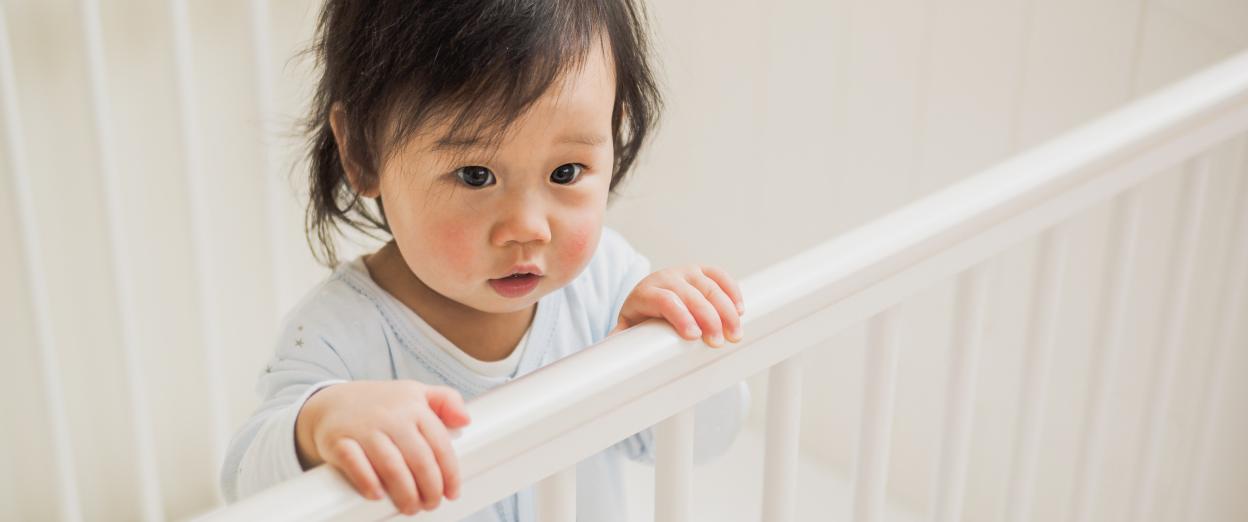 14month old toddler standing in crib. Baby is Asian. They look like they just woke up or have been crying with puffy under eyes and slightly messy dark hair