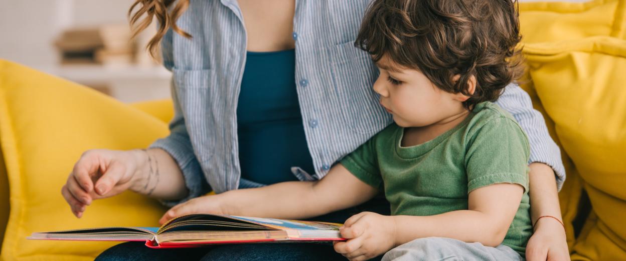 21 month old sitting on yellow couch looking at a book while sitting next to mom
