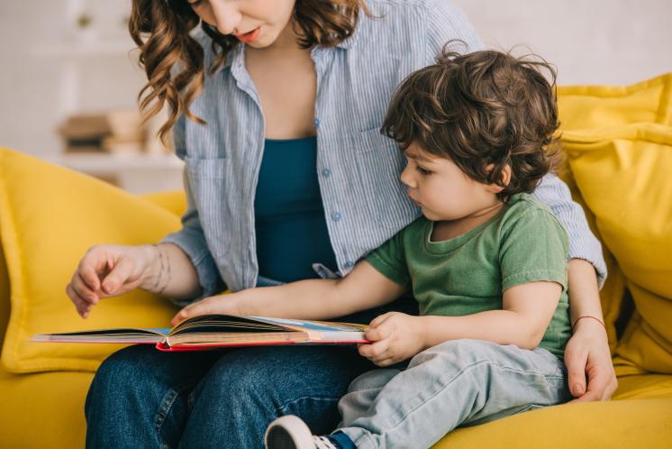 21 month old sitting on yellow couch looking at a book while sitting next to mom