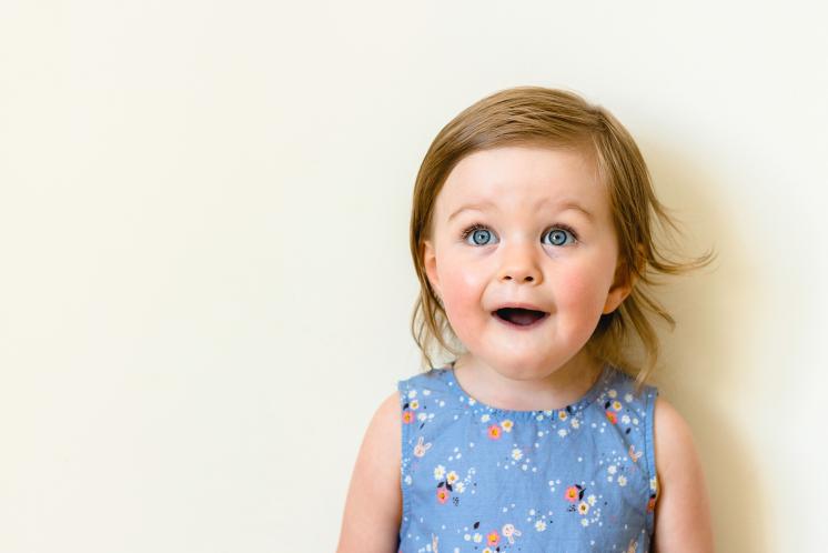 Older baby girl wearing blue tank top with light colored flowers looking up at the camera with a surprised face