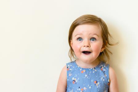 Older baby girl wearing blue tank top with light colored flowers looking up at the camera with a surprised face