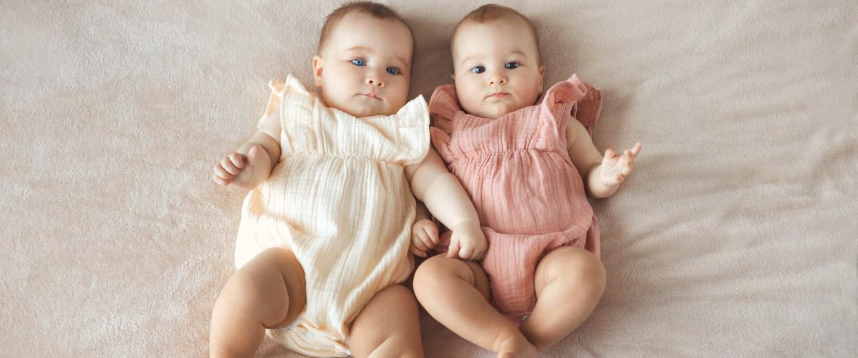 baby girl twins looking a the camera, one in off white the other in blush pink rompers