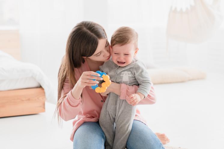 Mom kneeling down trying to calm overtired baby by shushing and holding out a blue and yellow rattle.