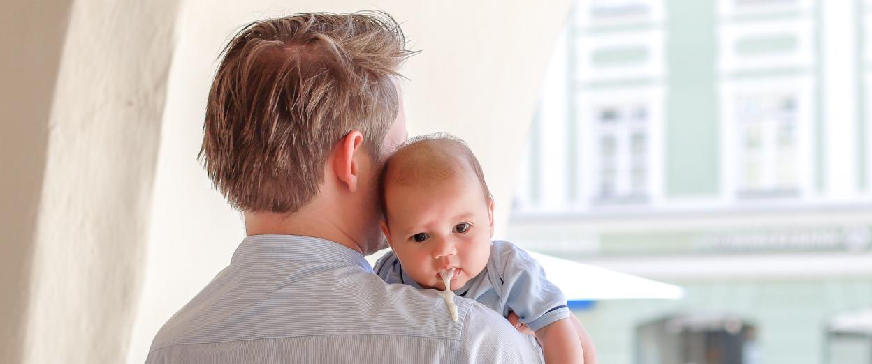 man facing away from camera with baby on his shoulder who is facing the camera as baby spits up over Dad's shoulder