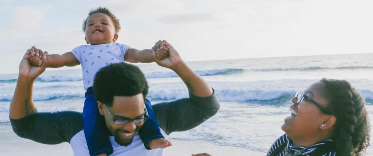 young family at the beach with baby on dad's shoulders, mom and dad smiling happily