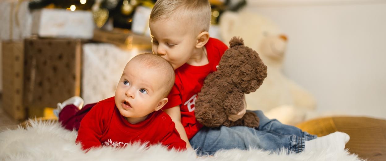 toddler brother kissing baby brother's head while sitting on the floor in front of the Christmas tree