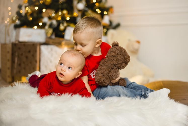 toddler brother kissing baby brother's head while sitting on the floor in front of the Christmas tree