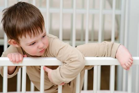 toddler in neutral pajamas lifting leg over the side of a white crib to climb out