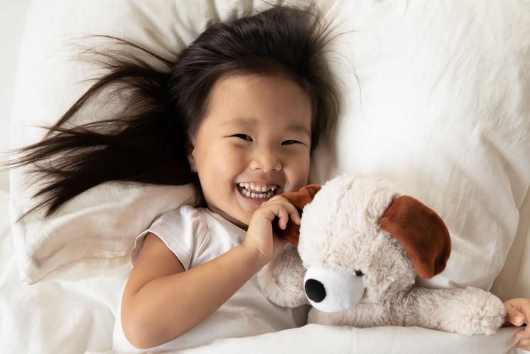 Toddler girl laying in white sheeted bed smiling at camera while holding a stuffed animal