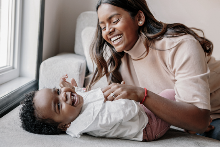17 month old playing with mom on couch smiling at the camera. Mom and baby have a dark skin tone. Baby has black curly hair and mom has dark brow hair. She is wearing a light beige mock neck shirt and a red bracelet.